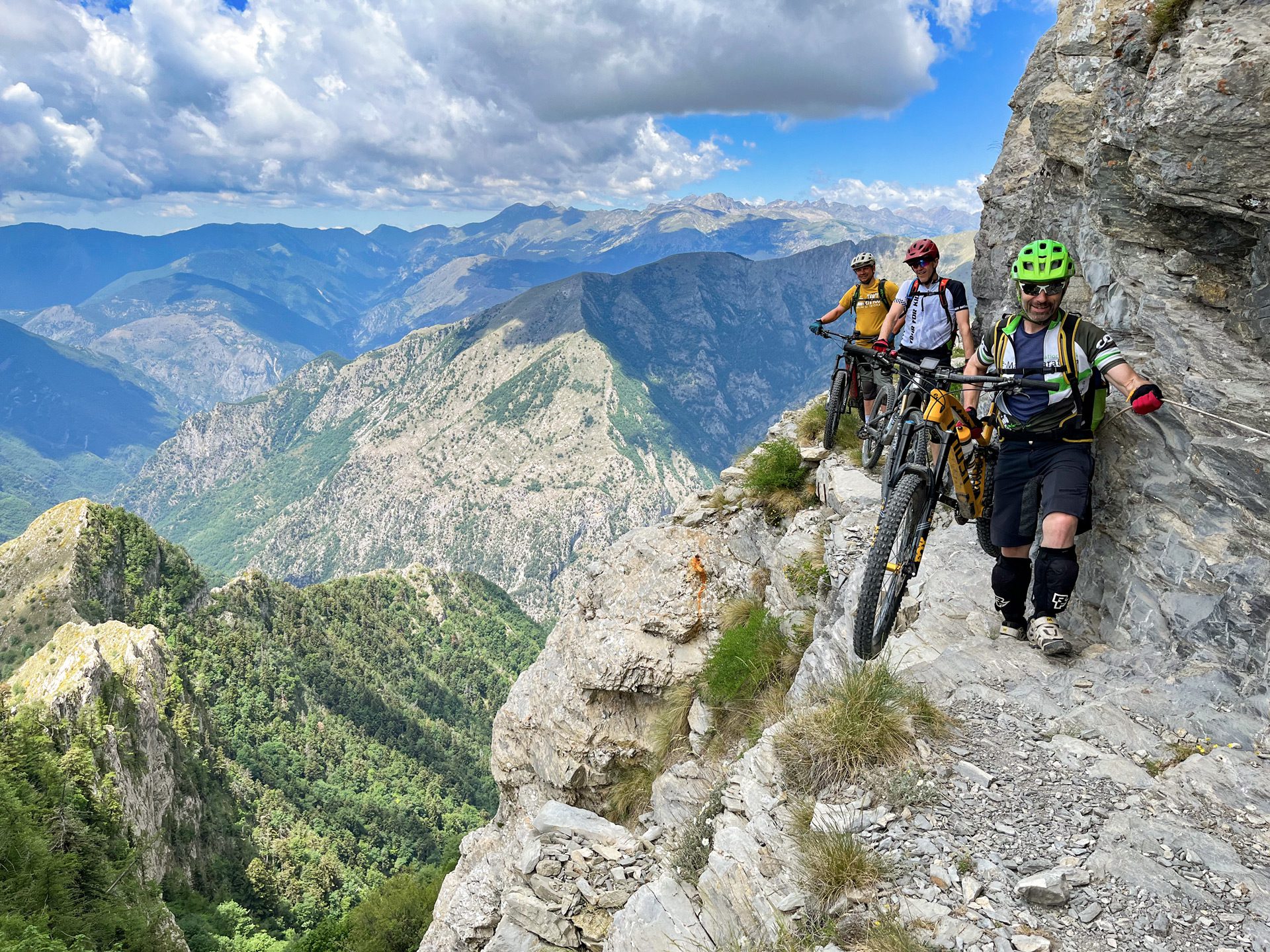 Berglandschaft; im Vordergrund rechts drei Mountainbiker, die ihre Räder am eine ausgesetzte Felskante entlangtragen.